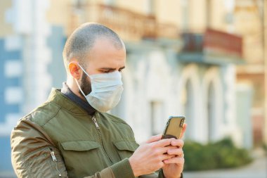 Akıllı telefon kullanan bir adam, şehir sokaklarında yayılan koronavirüsten kaçınmak için tıbbi maske takıyor. Yüzünde ameliyat maskesi olan bir adam COVID-19 'a karşı..