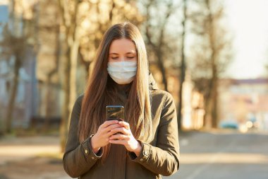 Akıllı telefon kullanan bir kadın, şehir sokaklarında yayılan koronavirüsten kaçınmak için tıbbi maske takıyor. Yüzünde ameliyat maskesi olan bir kız COVID-19 'a karşı..