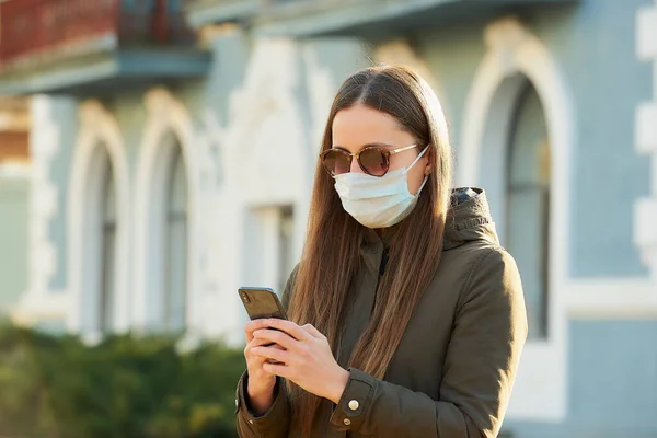 A girl using a smartphone wears a medical face mask to avoid the spread coronavirus on a city street. A woman in sunglasses with a surgical mask on the face against COVID-19.