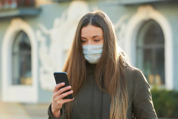 A woman using a smartphone wears a medical face mask to avoid the spread coronavirus on a city street. A close-up photo of a girl with a surgical mask on the face against COVID-19.
