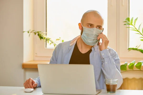 A man in a face mask working remotely on his laptop during the quarantine to avoid the spread coronavirus. A bald guy with a beard calling while working from home during the pandemic of COVID-19.