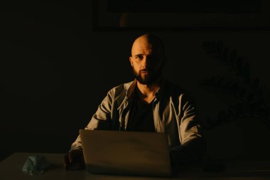 Sakallı bir adam karantina sırasında koronavirüsü yaymamak için bilgisayarını uzaktan kontrol ediyor. COVID-19 salgını sırasında akşamları evden çalışan bir işadamı.