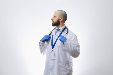 Bir doktor steteskop tutan tek kullanımlık tıbbi eldivenlerle poz verir. Sakallı kel bir doktor hastaları muayene etmeye hazırlanıyor..