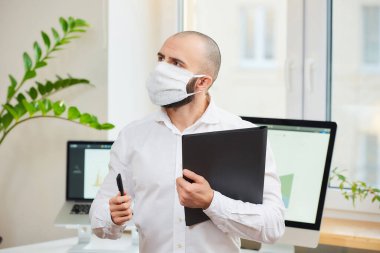 Coronavirus 'a karşı tıbbi maske takan bir adam (COVID-19). Elinde kalem ve klasörle arka planda bilgisayarları ve yeşil bitkileri olan bir yönetici çalışma alanına bakıyor. Coronavirus karantinası.