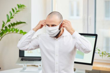 Gömlekli sakallı bir adam, Coronavirus 'a karşı tıbbi maske takıyor. İş yerinde bilgisayarları ve yeşil bitkileri olan bir mühendis. Coronavirus karantinası.