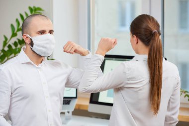 Koronavirüsün (SARS-CoV-2) yayılmasını önlemek için dirsek seği selamı. Tıbbi maskeli bir adam ve bir kadın ofiste çıplak elle buluşuyorlar. Sarılmak ya da tokalaşmak yerine, dirseklerini vururlar.