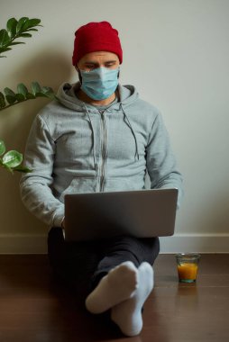 Karantina sırasında dizüstü bilgisayarı üzerinde çalışan tıbbi yüz maskesi takmış bir adam. COVID-19 salgını sırasında evde oturan bir programcı yerde çalışır..