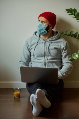 Karantina sırasında dizüstü bilgisayarı üzerinde çalışan tıbbi yüz maskesi takmış bir adam. COVID-19 salgını sırasında evde oturan bir işadamı yerde çalışıyor..