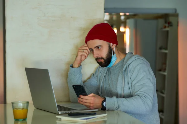 A man in the removed face mask working remotely on his laptop during the quarantine to avoid the spread coronavirus. A programmer works using a smartphone from home during the pandemic of COVID-19.