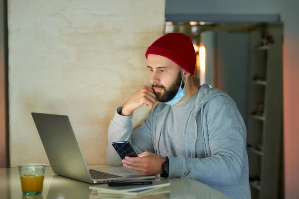 A man in the removed face mask working remotely on his laptop during the quarantine to avoid the spread coronavirus. A businessman works using a smartphone from home during the pandemic of COVID-19.