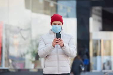 Koronavirüsten (COVID-19) korunmak için tıbbi yüz maskesi takan sakallı beyaz bir adam. Salgın yüzünden yüzünde cerrahi maske olan bir adam şehrin merkezinde akıllı telefon tutuyor..