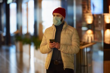 Koronavirüsten (COVID-19) korunmak için tıbbi yüz maskesi takan sakallı beyaz bir adam. Yüzünde cerrahi maske olan bir adam alışveriş merkezinde akıllı bir telefonla kalıyor..