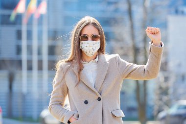 Paltolu bir kadın COVID-19 'un yayılmasını önlemek için tıbbi maske takıyor. Sokakta güneş gözlüklü bir kadın, Coronavirus 'a karşı verilen mücadelenin sembolü olarak yumruğunu gösteriyor..  