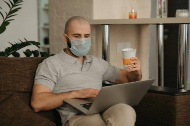 Tıbbi maske takan bir adam karantina sırasında koronavirüsü yaymamak için uzaktan kumandayla çalışıyor. Bir adam COVID-19 salgını sırasında elinde bir fincan kahve tutarak evden çalışıyor.