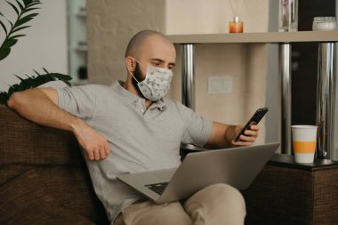 Uzaktan kumanda. Tıbbi maskeli bir adam karantina sırasında koronavirüsten kaçmak için bilgisayarını uzaktan kontrol ediyor. COVID-19 salgını sırasında elinde akıllı telefon tutan bir adam evde çalışıyor.