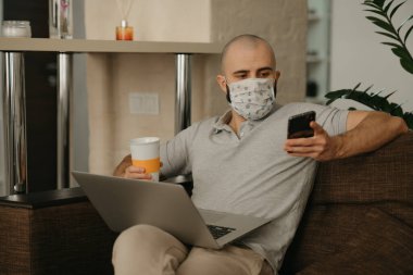 Yüzü maskeli bir adam karantina sırasında koronavirüsü yaymamak için uzaktan kumandayla çalışıyor. Bir adam COVID-19 salgını sırasında elinde telefon ve bir fincan kahveyle evde çalışıyor..