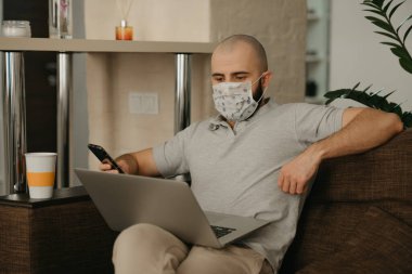 Uzaktan kumanda. Tıbbi maske takan bir adam karantina sırasında koronavirüsü yaymamak için uzaktan kumandayla çalışıyor. COVID-19 salgını sırasında evde çalışan bir adam elinde telefon tutuyor..