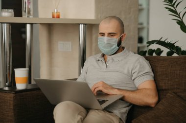 Uzaktan kumanda. Tıbbi maske takan bir adam karantina sırasında koronavirüsü yaymamak için uzaktan kumandayla çalışıyor. COVID-19 salgını sırasında evden çalışan sakallı bir adam..