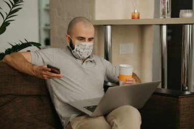 Yüzü maskeli bir adam karantina sırasında koronavirüsü yaymamak için uzaktan kumandayla çalışıyor. Bir adam COVID-19 salgını sırasında elinde telefon ve bir fincan çayla evde çalışıyor..