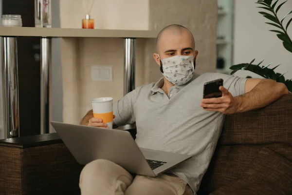 A man in a face mask working remotely on his laptop during the quarantine to avoid the spread coronavirus. A guy works from home holding a phone and a cup of coffee during the pandemic of COVID-19.