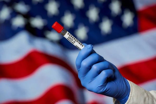 A vaccine against coronavirus. A hand of an infectious disease physician in a medical glove holding a test tube with a vaccine against COVID-19 against the background of the USA Stars and Stripes flag