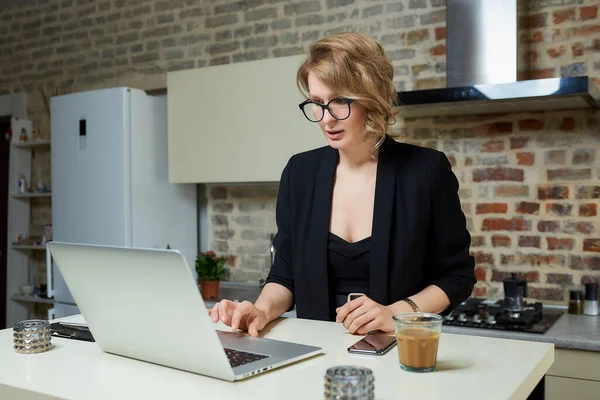 A woman in glasses works remotely on a laptop in her kitchen. A serious girl browsing news on the internet at home. A lady preparing for a lecture on a video call.