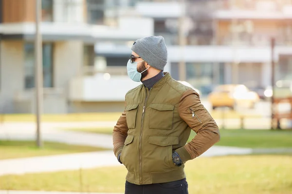 一个戴着医疗面罩的男人 以避免在舒适的街道上等待传播的考拉韦病毒 一个人把双手插在口袋里 戴着帽子 太阳镜 戴着口罩 戴着Covid — 图库照片