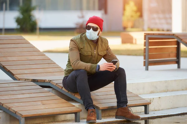 A man in a medical face mask to avoid the spread coronavirus holding a smartphone in the cozy street. A guy sitting on a wooden deck chair wears sunglasses and a face mask against COVID 19.