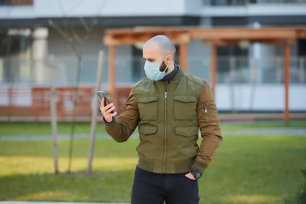 一个戴着医疗面罩的秃头男子在舒适的街道上检查他的智能手机 一个留着胡子的人侧着身子站着 戴着口罩对着Covid — 图库照片