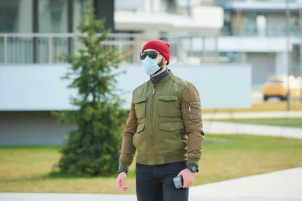 一个戴着医疗面罩的男人 为了避免在舒适的街上拿着智能手机而被传播开来 一个拿着红帽子 太阳镜和防毒面具等着我们的人 — 图库照片