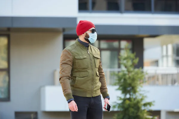 一个戴着医疗面罩的男人 为了避免在舒适的街上拿着智能手机而被传播开来 一个人四处张望 头戴红帽子 戴着太阳镜 还戴着防毒面具 — 图库照片
