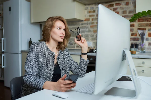 A woman with glasses works remotely on a desktop computer in her studio. A lady uses a phone during a video conference at home. A female professor listening to student\'s answers on an online lesson.