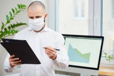 Coronavirus 'a karşı tıbbi maske takan bir adam (COVID-19). Çalışma alanında elinde kalem ve dosya tutan bir ofis çalışanı. Arka planda bilgisayarlar ve yeşil bitkiler var. Coronavirus karantinası