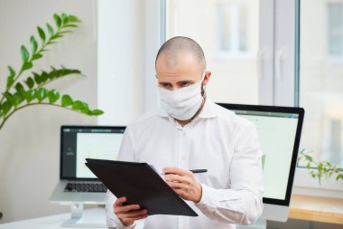 Coronavirus 'a karşı tıbbi maske takan bir adam (COVID-19). Bir ofis çalışanı çalışma alanında bilgisayarlarla ve arka planda yeşil bitkilerle notlar alıyor. Coronavirus karantinası