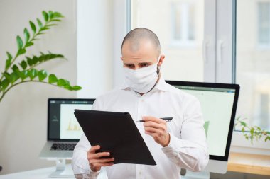 Coronavirus 'a karşı tıbbi maske takan bir adam (COVID-19). Arka planda bilgisayarlar ve yeşil bitkilerle çalışma alanında notlar alan bir yönetici. Coronavirus karantinası.