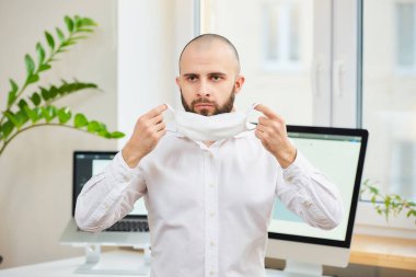 Gömlekli sakallı bir adam, Coronavirus 'a karşı tıbbi maske takıyor. Çalışma alanında bilgisayarları ve arka planda yeşil bitkileri olan bir yönetici. Coronavirus karantinası.
