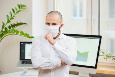 Tıbbi yüz maskesi takmış bir adam Coronavirus 'a karşı (COVID-19) yumruğuna öksürüyor. Çalışma alanında bilgisayarları ve arka planda yeşil bitkileri olan bir yönetici. Coronavirus karantinası.
