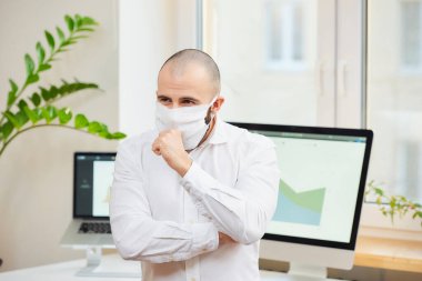 Tıbbi yüz maskesi takmış bir adam Coronavirus 'a karşı (COVID-19) güçlü bir şekilde yumruğuna öksürüyor. Çalışma alanında bilgisayarları ve arka planda yeşil bitkileri olan bir yönetici. Coronavirus karantinası.
