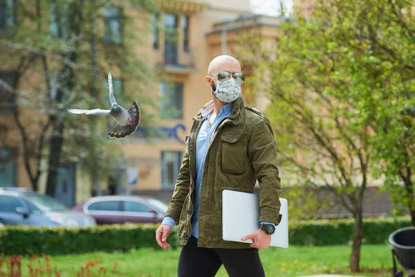 A bald man in a medical face mask to avoid the spread coronavirus walks with a laptop in the park. A guy wears n95 face mask and a pilot sunglasses on the street near a flying pigeon.