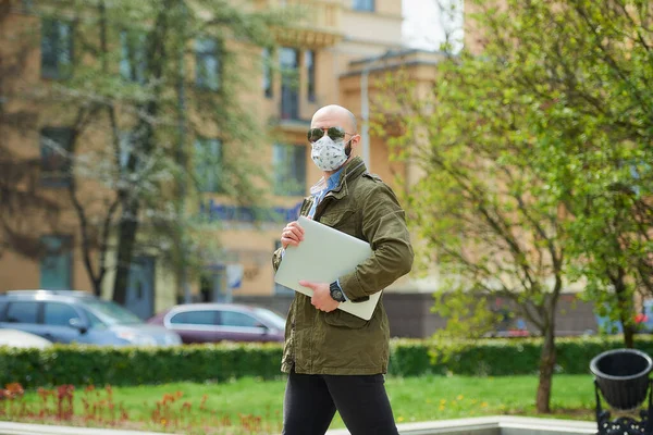 A bald man with a beard in a medical face mask to avoid the spread coronavirus walks with a laptop in the park. A guy wears n95 face mask and a pilot sunglasses on the street of the city.