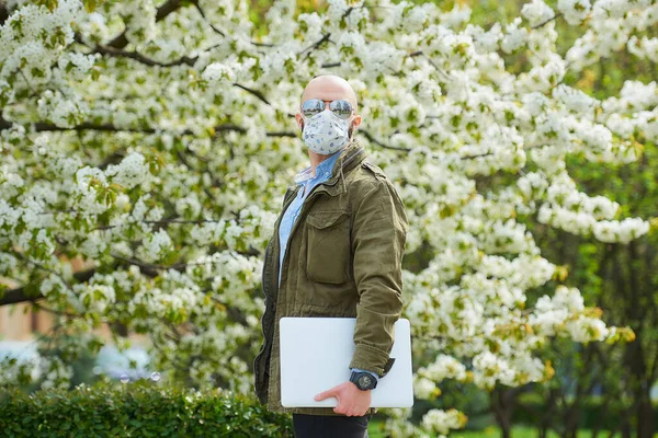 A bald man with a beard in a medical face mask to avoid the spread coronavirus walks with a laptop in the park. A guy wears n95 face mask and a pilot sunglasses in the garden between flowering trees.