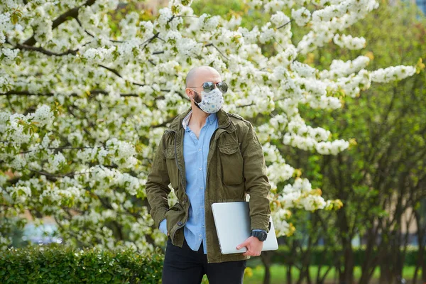 一个秃头的男人 戴着医疗面罩 留着胡子 以避免在公园里带着笔记本电脑散步 一个戴着N95口罩 戴着飞行员太阳镜的男人出现在花木丛中的花园里 — 图库照片