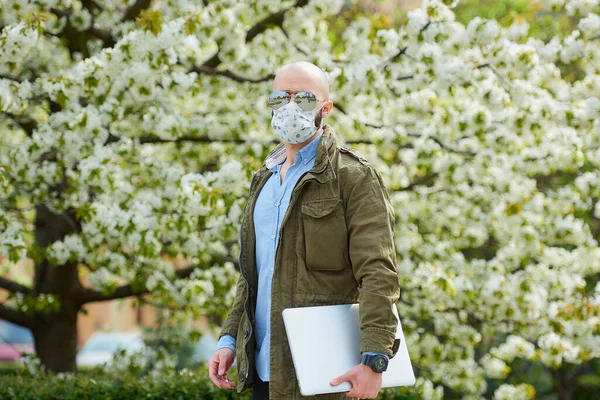 A bald man with a beard in a medical face mask to avoid the spread coronavirus walks with a laptop in the park. A guy wears n95 face mask and a pilot sunglasses in the garden between flowering trees.