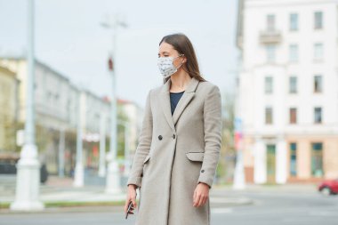 Tıbbi maske takmış uzun saçlı esmer bir kız koronavirüsten kaçınmak için sokakta akıllı telefon tutuyor. COVID-19 'a karşı maske takan bir kadın şehirde başka bir yere bakıyor..