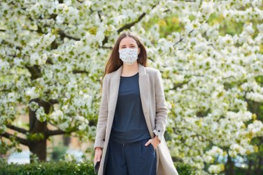 Koronavirüsten korunmak için tıbbi maske takan bir kız parkta akıllı telefon tutuyor. COVID-19 'a karşı maskeli bir kadın çiçek açan ağaçlar arasındaki sosyal mesafeyi koruyor..
