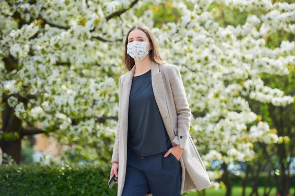 Koronavirüsten korunmak için tıbbi maske takan bir kız parkta akıllı telefon tutuyor. COVID-19 'a karşı maskeli bir kadın çiçek açan ağaçlar arasındaki sosyal mesafeyi koruyor..