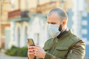 Akıllı telefon kullanan bir adam, şehir sokaklarında yayılan koronavirüsten kaçınmak için tıbbi maske takıyor. Yüzünde ameliyat maskesi olan bir adam COVID-19 'a karşı..