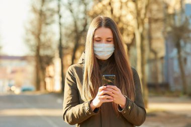 Akıllı telefon kullanan bir kadın, şehir sokaklarında yayılan koronavirüsten kaçınmak için tıbbi maske takıyor. Yüzünde ameliyat maskesi olan bir kız COVID-19 'a karşı..