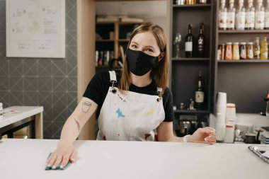 Siyah maskeli, dövmeli bir bayan barista gülümsüyor ve bir kahve dükkanında tezgahı temizliyor. Güzel bir garson kafeyi dezenfekte ediyor..