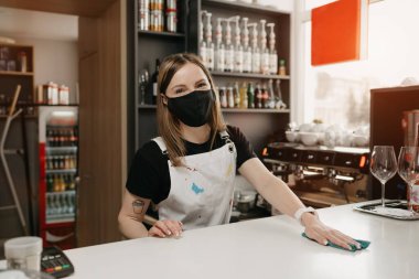 Siyah maskeli, dövmeli bir bayan barista gülümsüyor ve bir kahve dükkanında tezgahı temizliyor. Güzel bir garson müşterileri beklerken bir kafeyi dezenfekte ediyor..